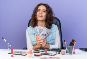 pleased pretty Caucasia woman sitting table with makeup tools holding money