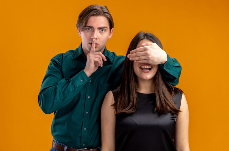 young couple valentines day strict guy covered girl eyes with hand showing silence gesture isolated orange background