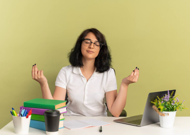 young pleased pretty caucasian schoolgirl wearing glasses sits desk with school tools pretends meditate with raised hands isolated green space with copy space